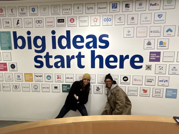 two women posing in front of their logo on the wall